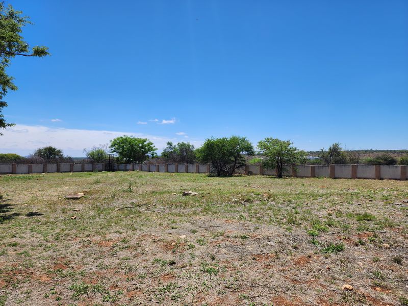 A view of a vacant residential plot featuring a perimeter wall and natural vegetation under a clear blue sky.