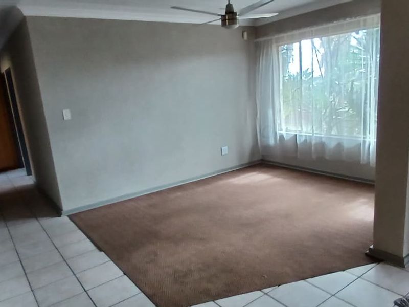 An empty living room featuring a combination of tiled and carpeted flooring, a ceiling fan, and a wooden door leading to a hallway.