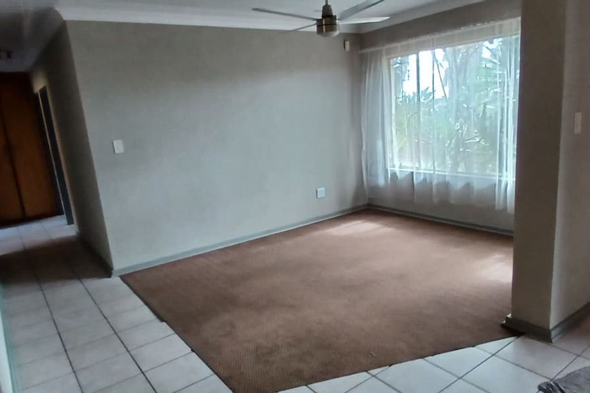 An empty living room featuring a combination of tiled and carpeted flooring, a ceiling fan, and a wooden door leading to a hallway.