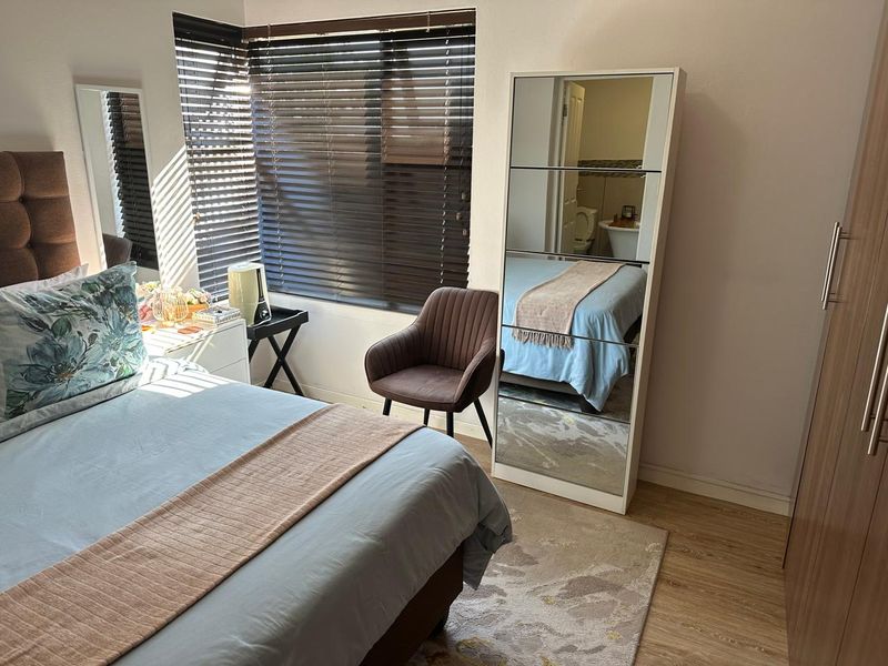 A well-maintained bedroom featuring laminate flooring, venetian blinds, and a built-in wardrobe, with an en-suite bathroom visible in the mirror reflection.