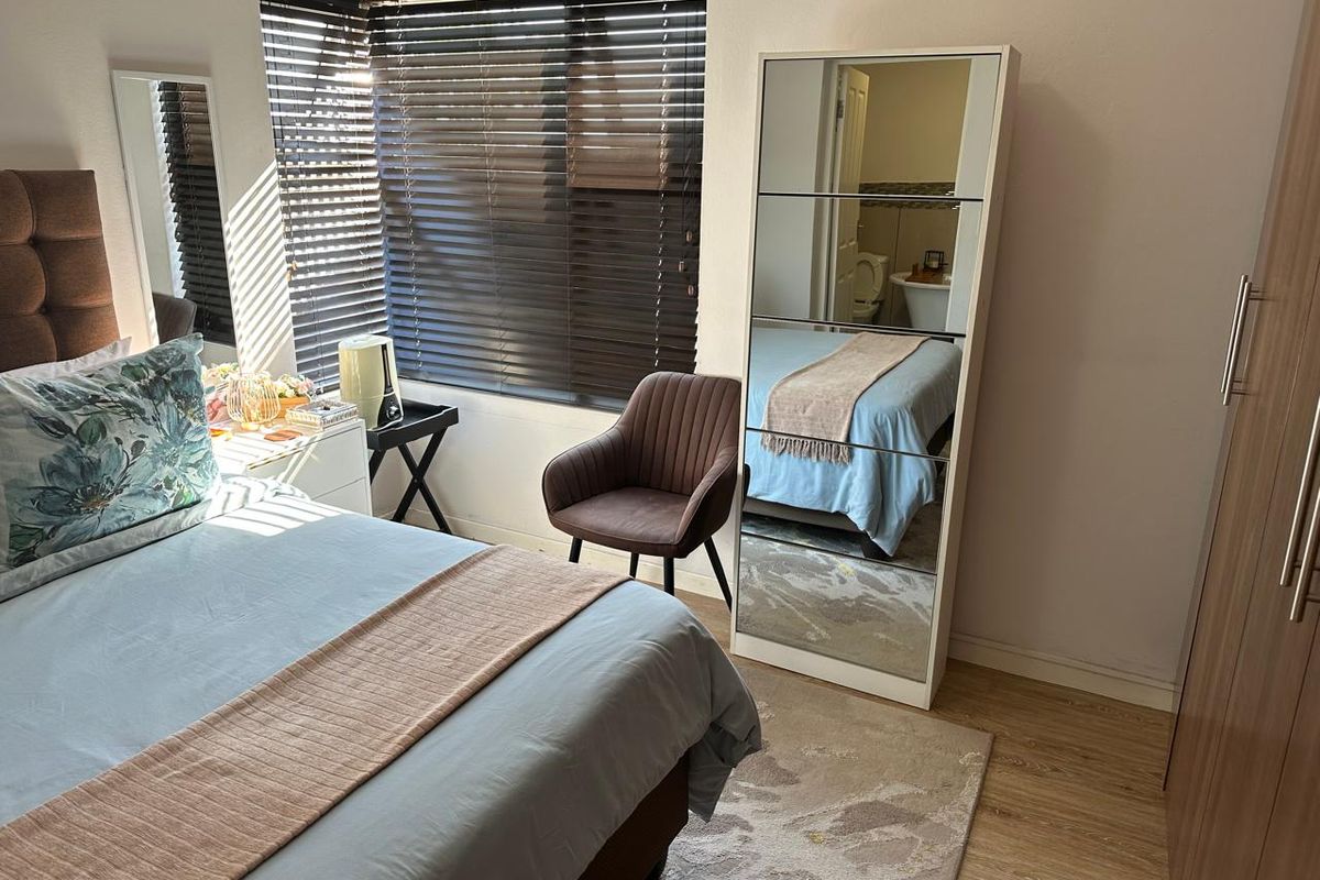 A well-maintained bedroom featuring laminate flooring, venetian blinds, and a built-in wardrobe, with an en-suite bathroom visible in the mirror reflection.