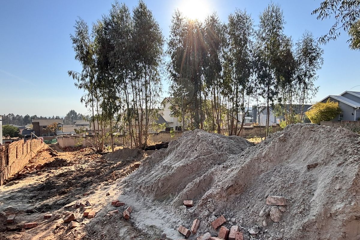 A rear outdoor area currently undergoing site preparation, featuring exposed soil, construction debris, and established trees against a boundary wall.