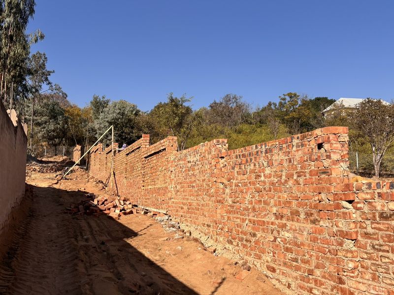 An exterior view showing an unfinished brick boundary wall along a dirt driveway in a rural or semi-rural setting.