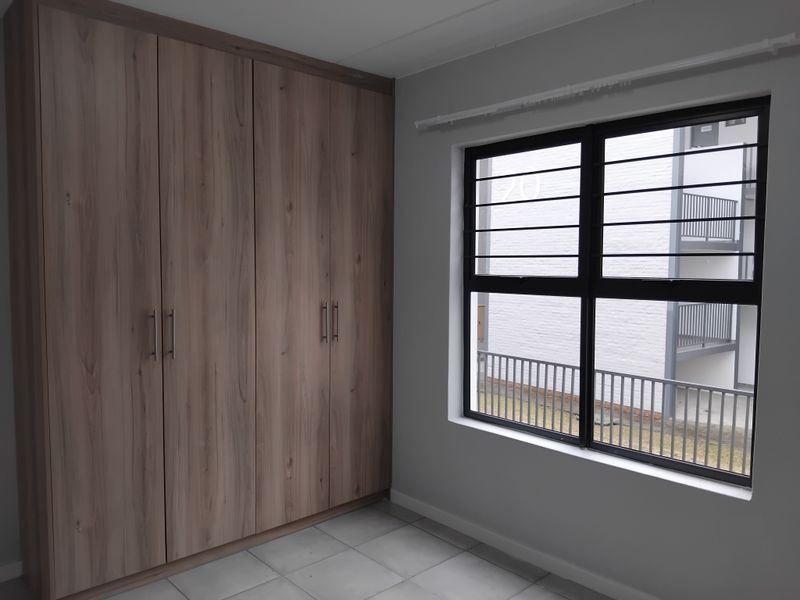 A clean, empty bedroom featuring modern wood-finish built-in cupboards, tiled flooring, and a window fitted with security bars.