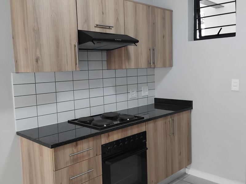 A clean, modern kitchen featuring light wood-finish cabinetry, black countertops, a built-in oven and hob, and a white tiled backsplash.