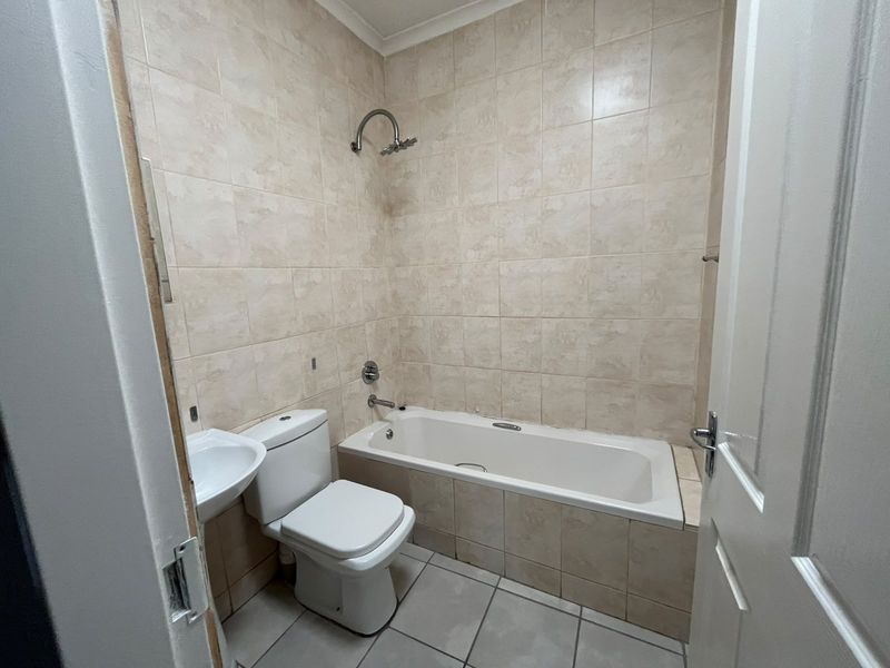 A functional bathroom featuring a bathtub with shower head, toilet, and wall-mounted basin, finished with neutral wall and floor tiles.
