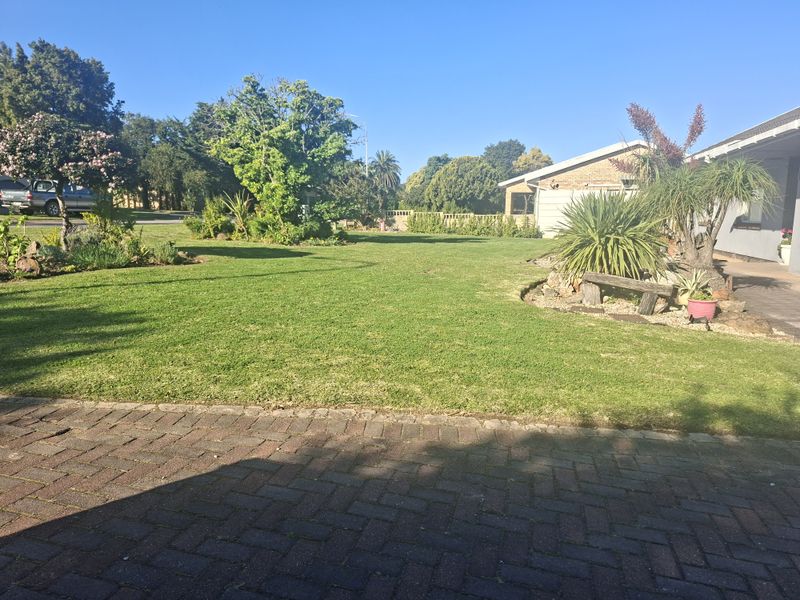 A well-maintained front garden featuring a spacious green lawn, established trees, and neat brick paving leading to the property entrance.