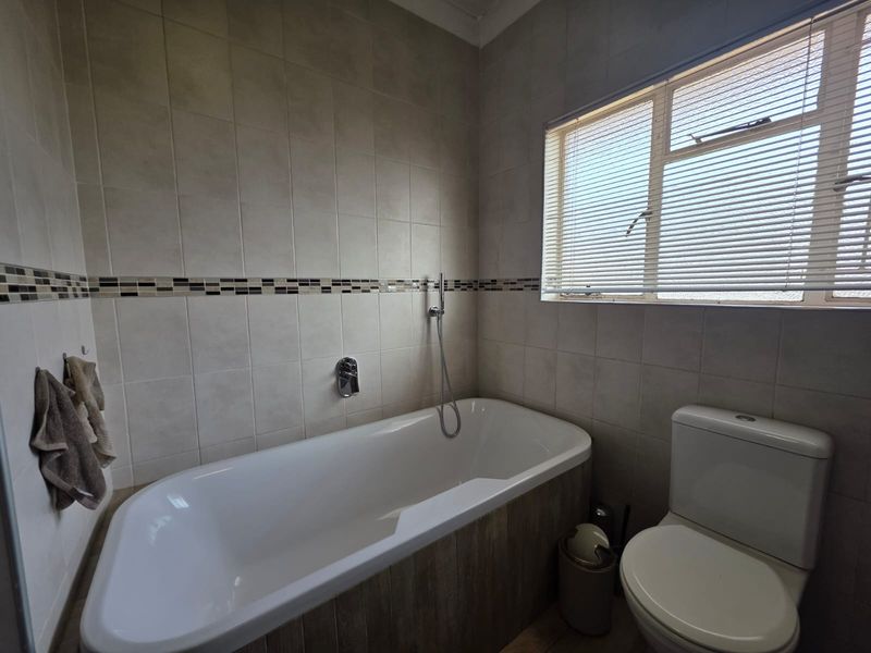 A functional bathroom featuring a built-in bathtub with wall-mounted mixer, ceramic wall tiling with a decorative border, and a standard dual-flush toilet.