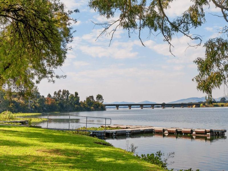 Scenic waterfront view featuring a floating jetty with a metal handrail, overlooking a calm lake with a bridge in the distance.
