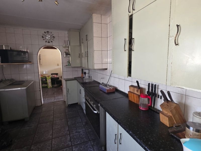 A dated kitchen featuring original cabinetry, tiled backsplash, and dark tiled flooring, leading through an archway to an adjacent room.
