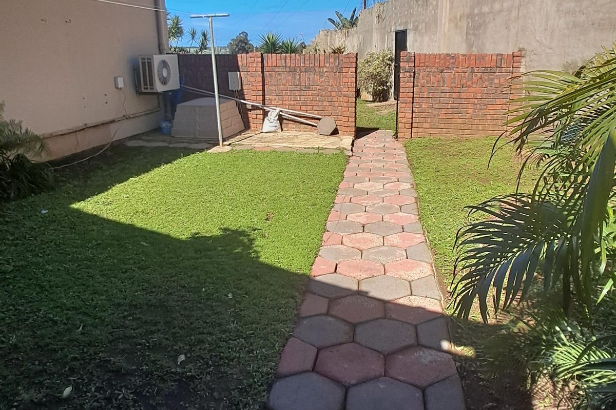 A rear garden area featuring a hexagonal paved walkway, a lawn, and a brick boundary wall with an external air conditioning unit.
