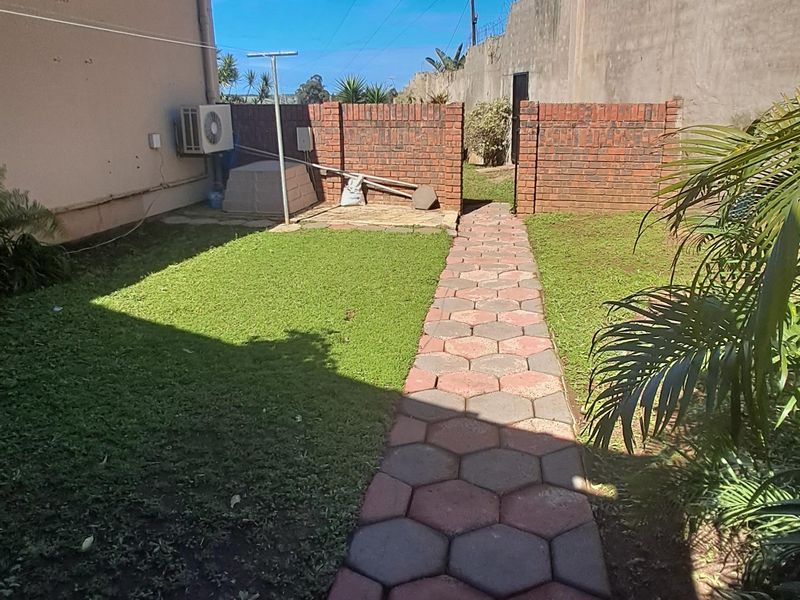 A rear garden area featuring a hexagonal paved walkway, a lawn, and a brick boundary wall with an external air conditioning unit.