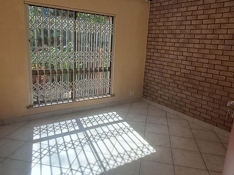 A bright, empty bedroom featuring neutral tiled flooring, an exposed brick feature wall, and a window fitted with a security gate.