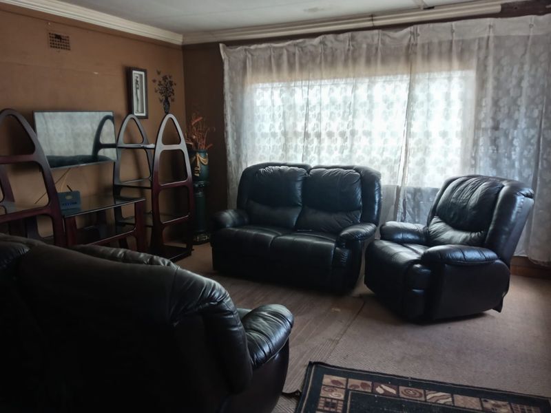 A living room featuring a large window with curtains, carpeted flooring, and coved ceiling details, offering a dated but functional space.