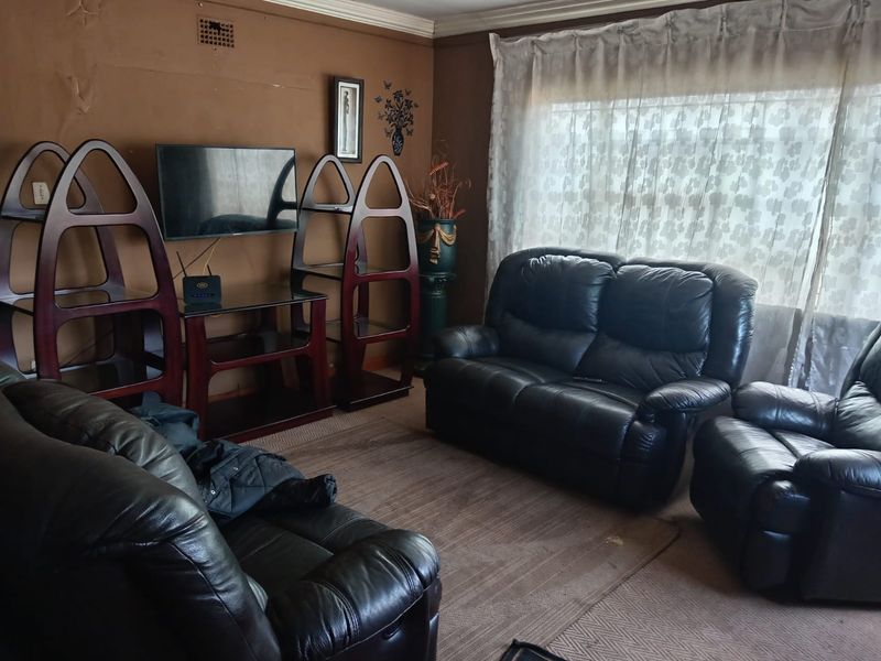 A living room featuring brown walls with visible surface wear and peeling paint, large window with curtains, and a ventilation grille.