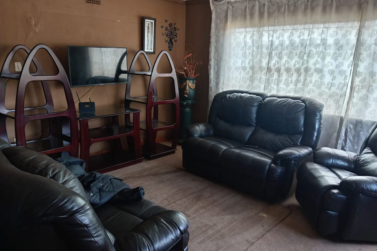 A living room featuring brown walls with visible surface wear and peeling paint, large window with curtains, and a ventilation grille.