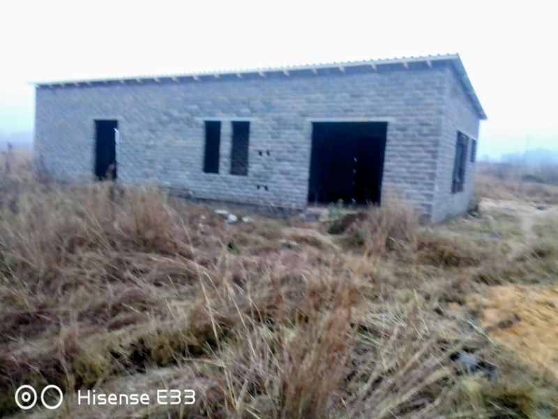 An unfinished single-story residential structure featuring exposed brick walls and a metal roof, set on an undeveloped plot of land.