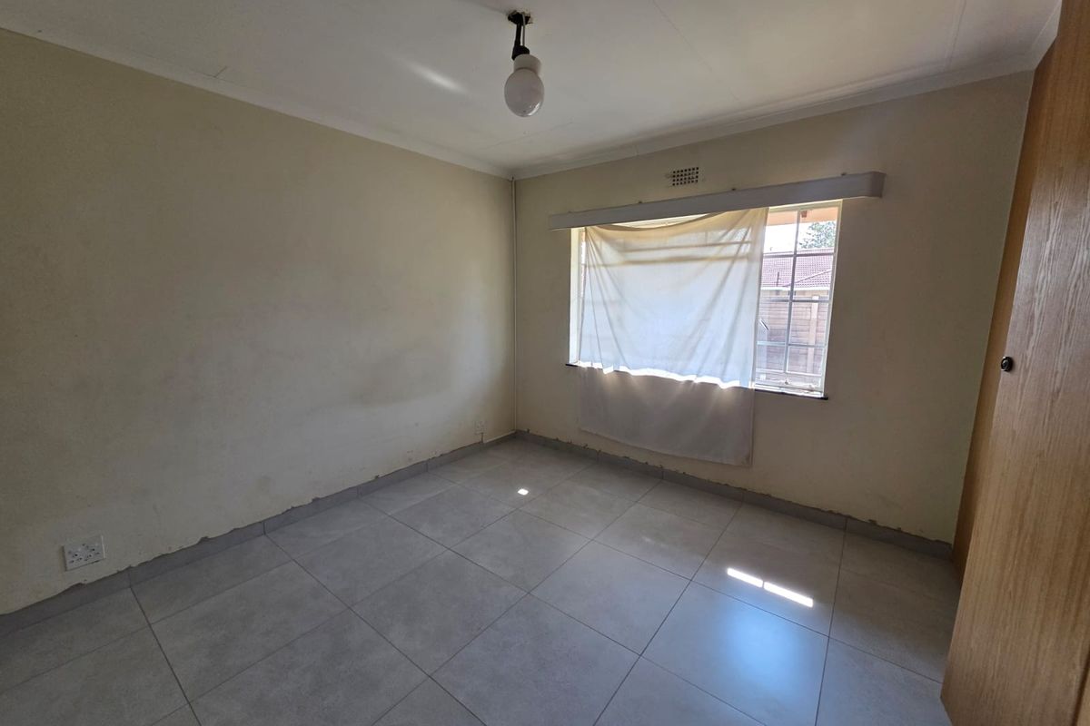 An empty bedroom featuring tiled flooring, a built-in cupboard, and a window with natural light. The room requires minor cosmetic updates to skirting and paint.
