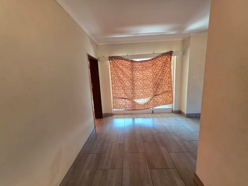 An empty living area featuring neutral tiled flooring, cove cornicing, and a large window with a curtain.
