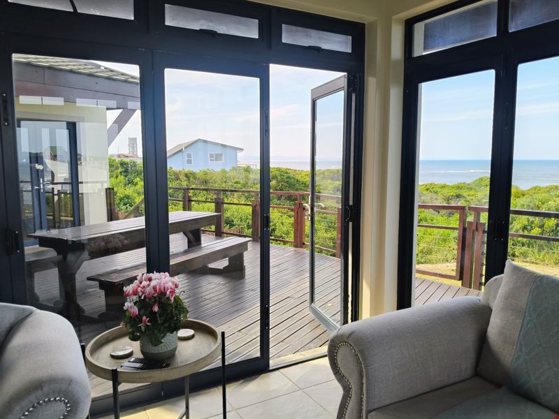 A bright living area featuring large black-framed glass doors that open onto a spacious timber deck with scenic coastal views.