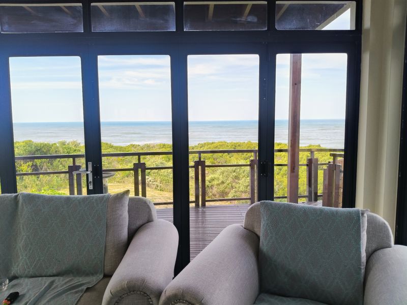A bright living area featuring large black aluminium stacker doors that open onto a timber deck with expansive ocean and coastal views.