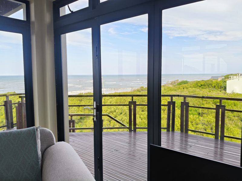 A scenic view of the ocean and coastal vegetation captured through large glass doors leading onto a timber deck with a metal balustrade.