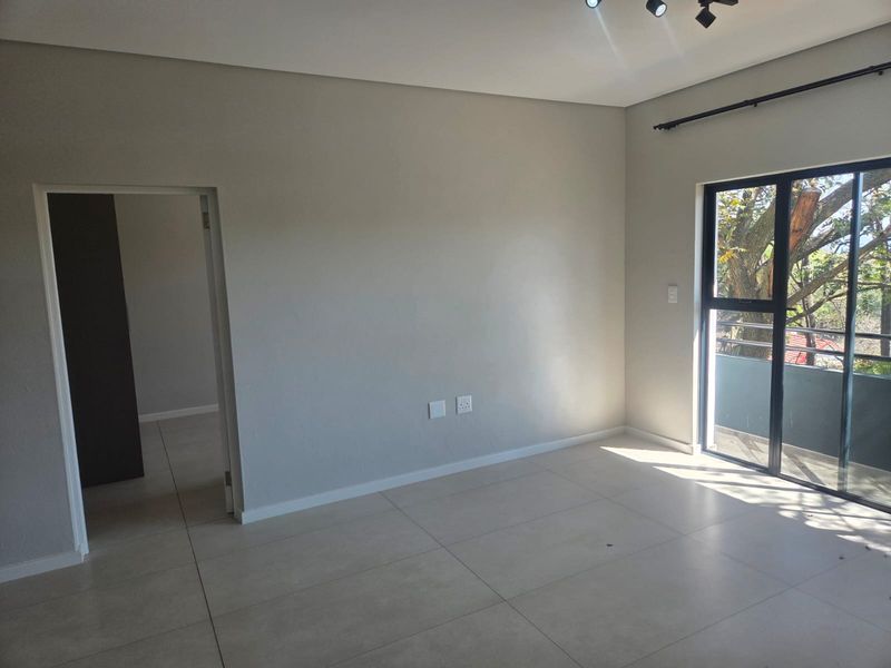 A bright, empty living room featuring neutral tiled flooring, modern track lighting, and a sliding glass door leading to an outdoor balcony.