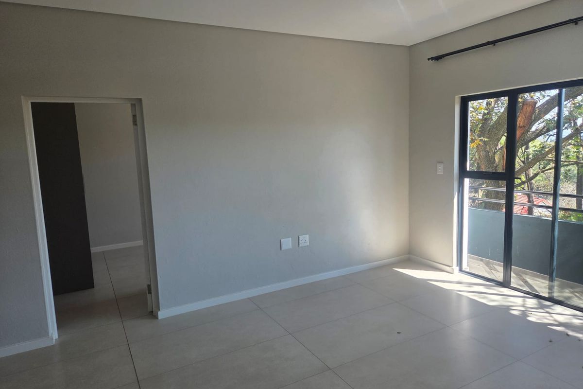 A bright, empty living room featuring neutral tiled flooring, modern track lighting, and a sliding glass door leading to an outdoor balcony.