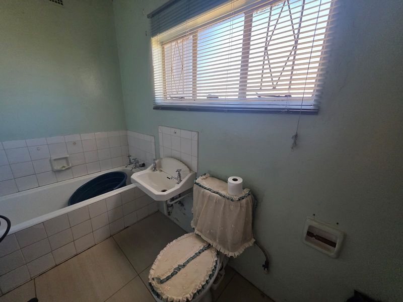 A dated bathroom featuring a built-in bathtub, wall-mounted basin, and toilet, with tiled splashbacks and flooring, requiring cosmetic updates to modernize the space.