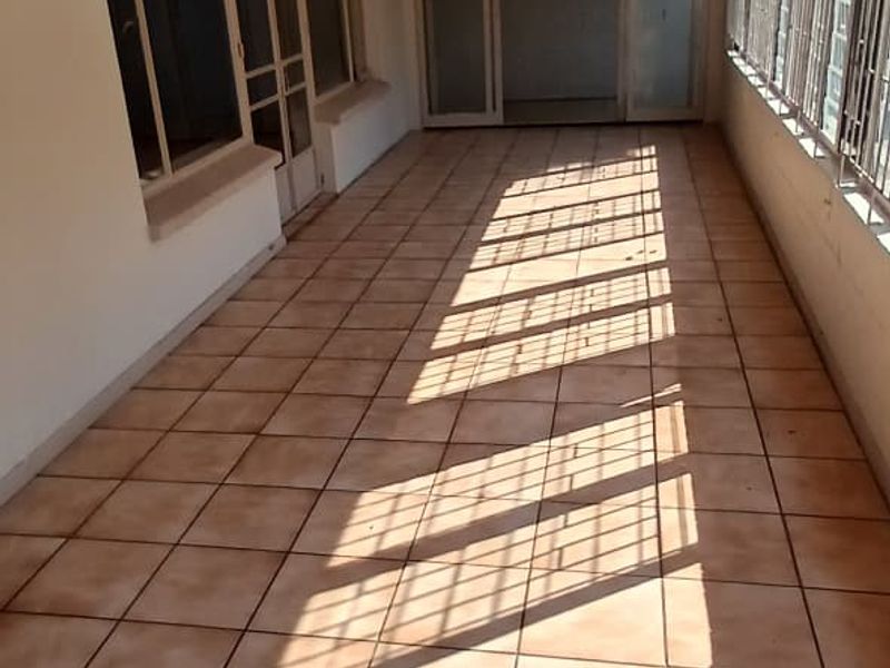 A spacious, enclosed patio area featuring tiled flooring, exposed timber ceiling beams, and large sliding glass doors with security bars.