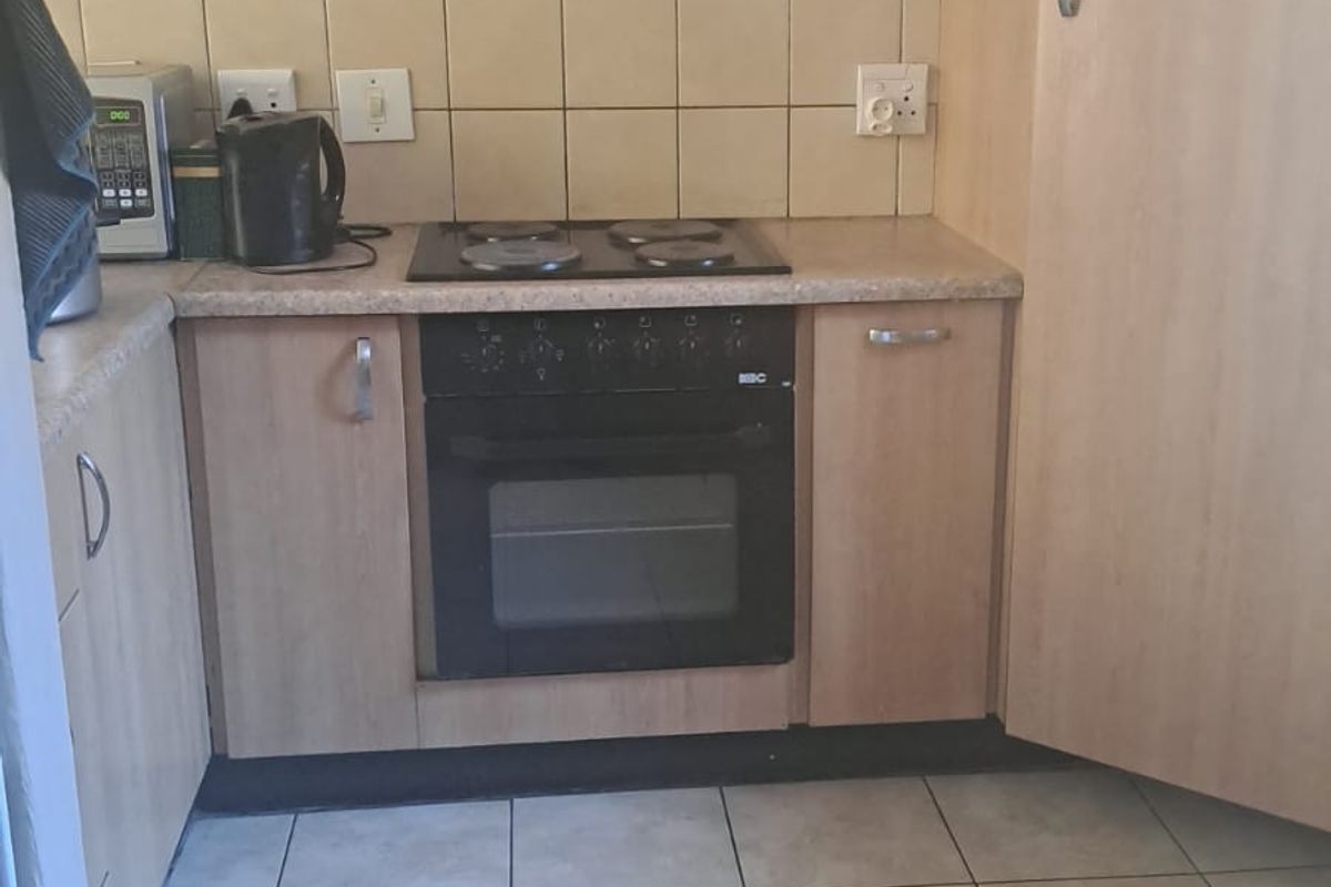 A compact kitchen featuring light wood-look cabinetry, a built-in oven with electric hob, tiled backsplash, and neutral ceramic floor tiles.