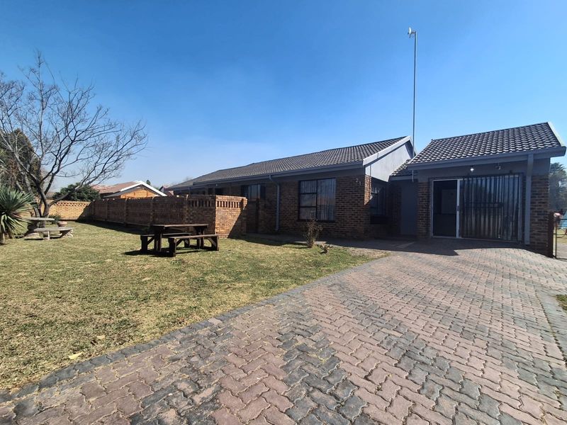 Front view of a single-story brick house featuring a paved driveway, tiled roof, and a surrounding brick boundary wall with a lawn area.