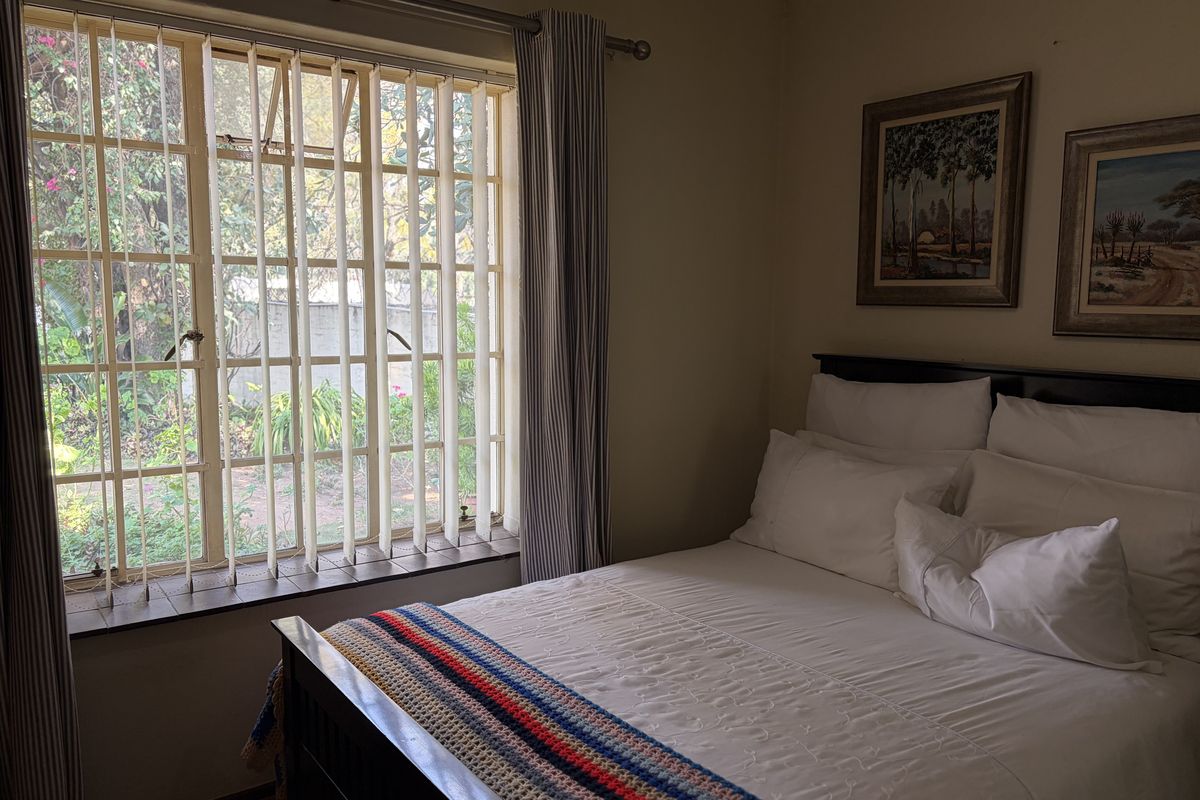 A cozy bedroom featuring a large window with security bars, vertical blinds, and neutral wall finishes, offering a view of the garden.