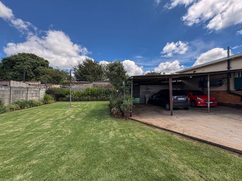Spacious rear garden featuring a large lawn area, a concrete carport, and a secure precast concrete boundary wall.