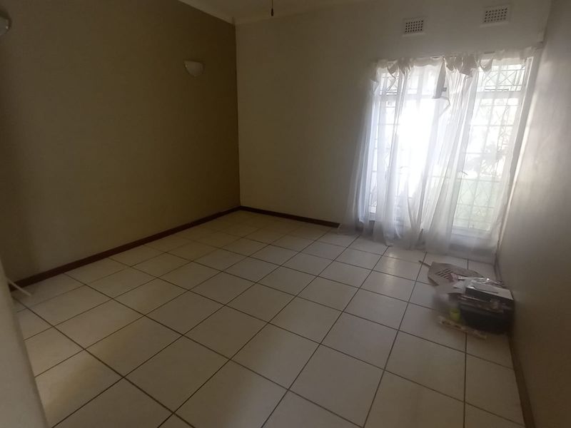 An empty living room featuring neutral tiled flooring, a ceiling fan, and a sliding glass door with security gate providing natural light.