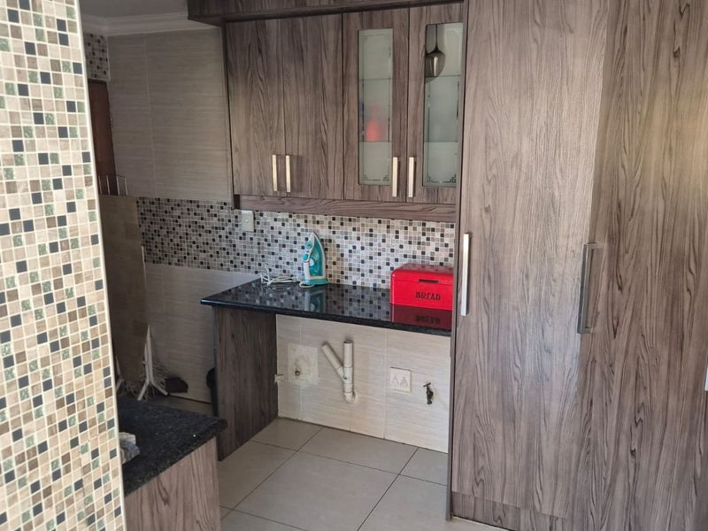A functional scullery area featuring wood-look cabinetry, granite countertops, and mosaic tile backsplash with dedicated plumbing connections for appliances.
