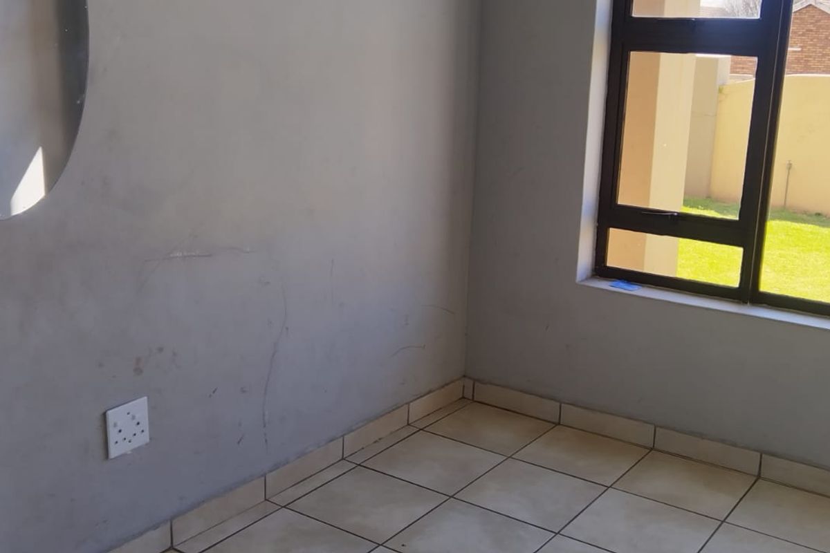 An empty bedroom featuring tiled flooring and an aluminium window frame, showing signs of wear on the wall surfaces.