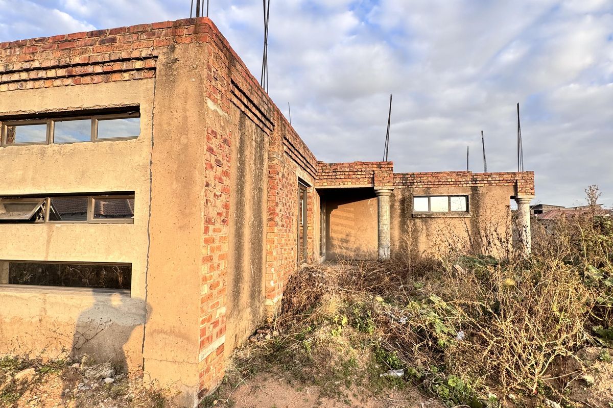 An unfinished residential structure featuring exposed brickwork, concrete columns, and horizontal window frames, set on a plot with overgrown vegetation.