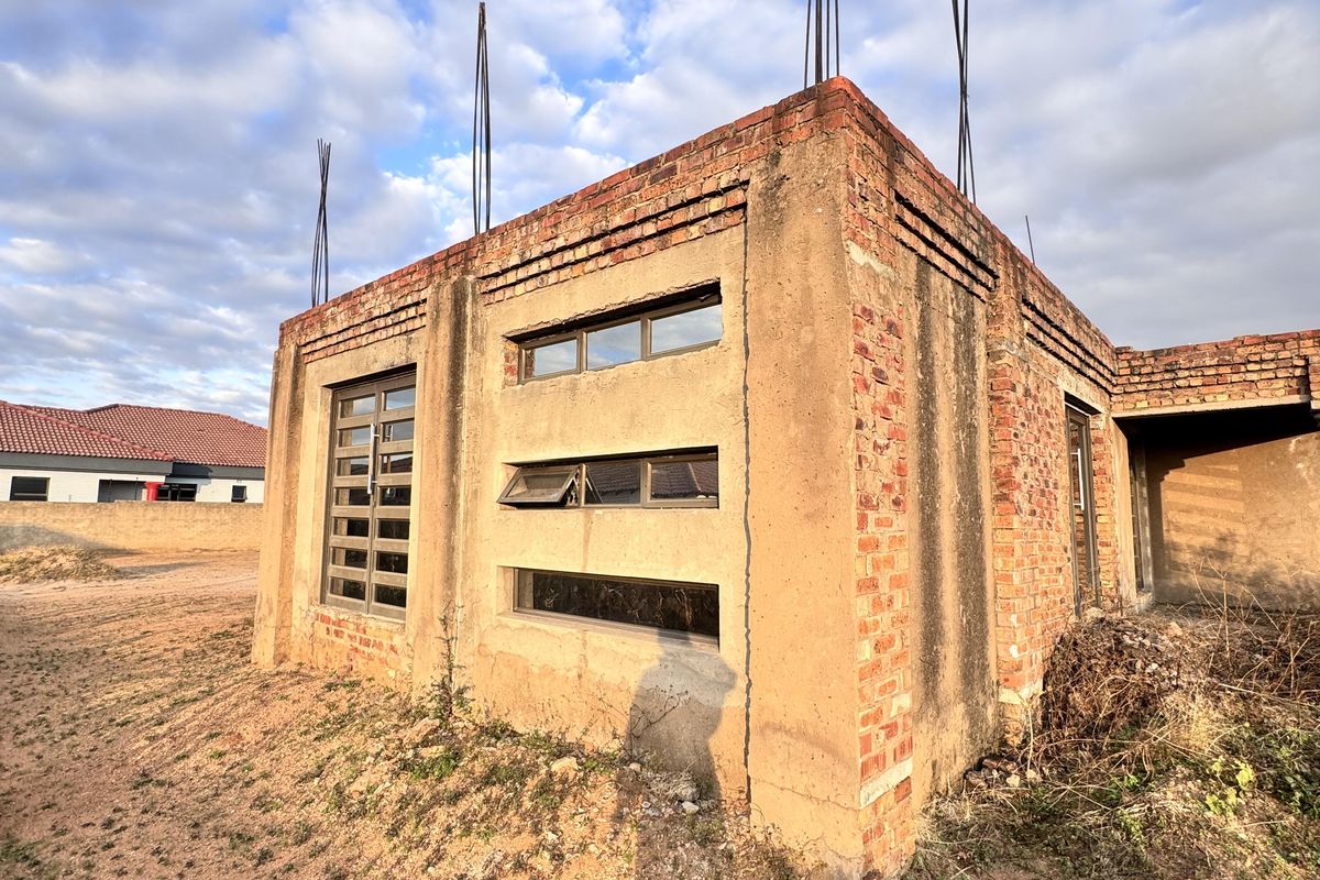An unfinished residential structure showing exposed brickwork, aluminium window frames, and protruding rebar, indicating a property currently under construction.