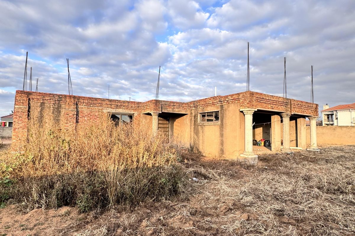 An unfinished residential structure showing exposed brickwork, concrete columns, and protruding rebar, set on a large plot requiring significant completion and landscaping.