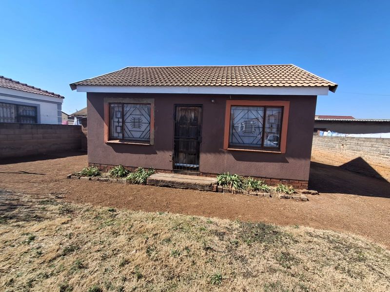 A detached single-story house featuring a tiled roof, security gates, and burglar bars, set within a fenced plot with a basic garden area.