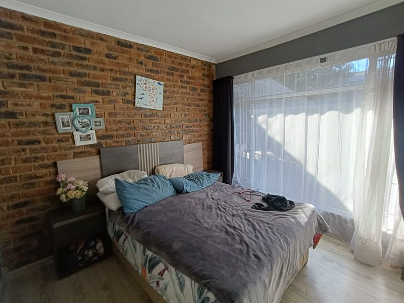 A well-lit bedroom featuring a rustic exposed brick feature wall, modern laminate flooring, and large windows allowing for ample natural light.