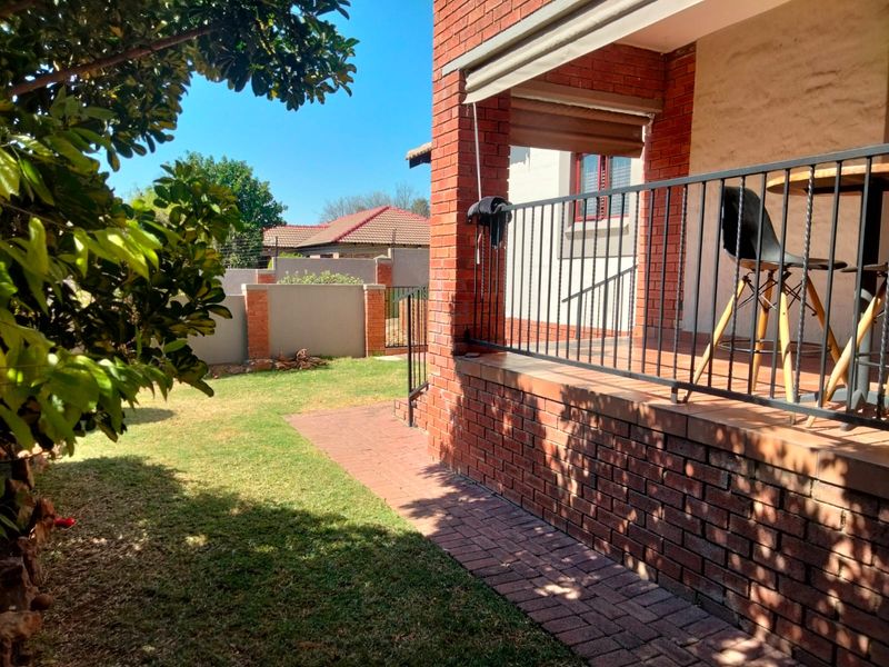 A covered brick patio area featuring a metal railing, retractable awning, and direct access to the surrounding garden and paved walkway.