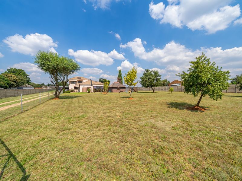 Expansive garden area featuring mature trees, perimeter fencing, a concrete boundary wall, and a small thatched lapa structure in the distance.