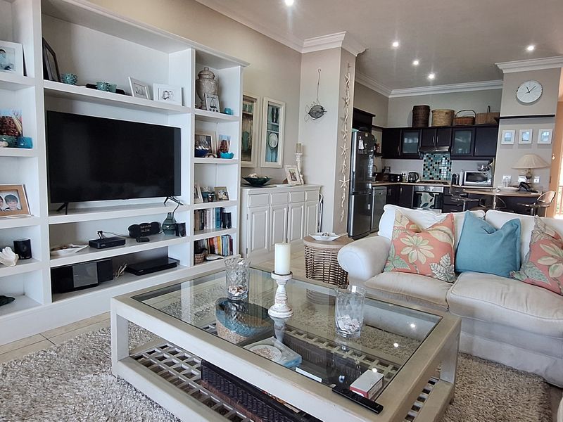Spacious open-plan living area featuring a large built-in media shelving unit, recessed lighting, and a functional kitchen with dark cabinetry.