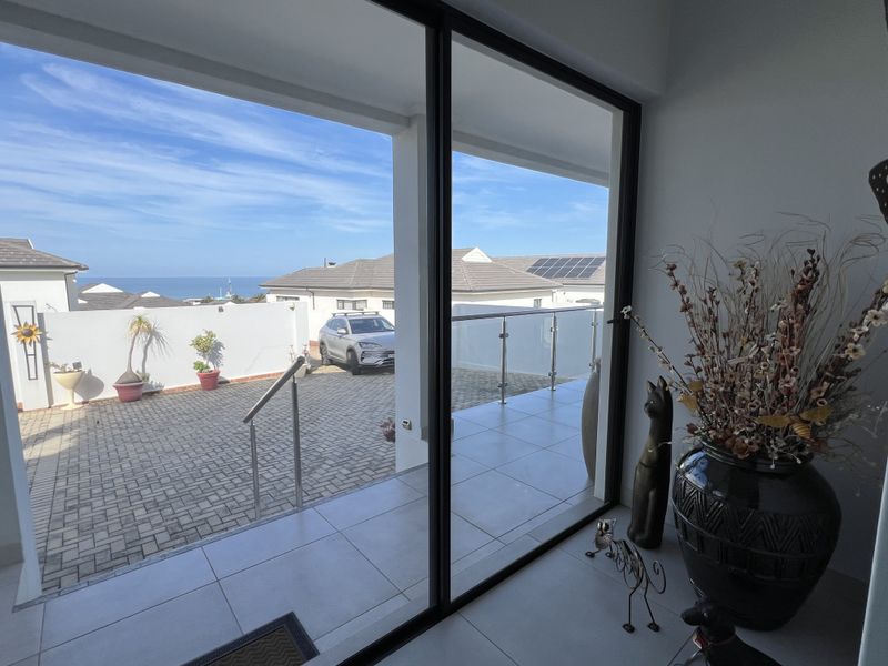A covered patio area featuring large sliding glass doors, tiled flooring, and a glass balustrade, overlooking a paved driveway and distant ocean views.