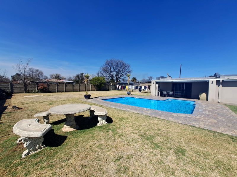 A spacious backyard featuring a swimming pool with paved surround, a covered patio area, and a large lawn enclosed by perimeter fencing.