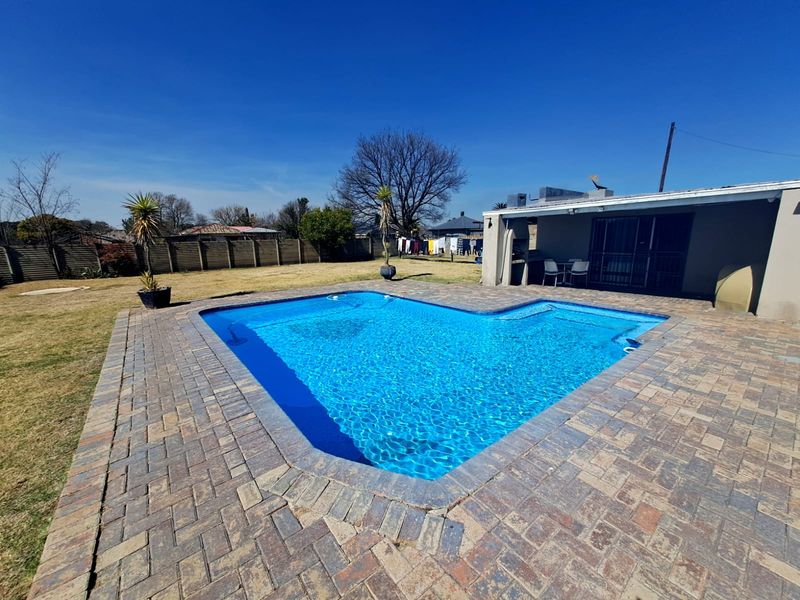 A well-maintained swimming pool area featuring a paved deck and an adjacent covered patio, set within a spacious backyard with perimeter fencing.