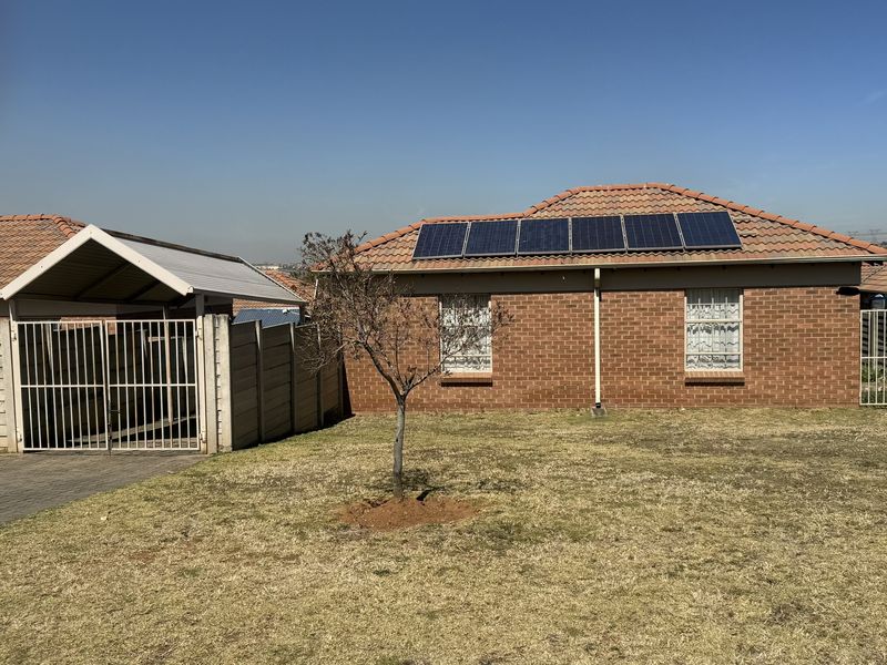 Rear view of a brick house featuring a tiled roof with installed solar panels, a small garden, and an adjacent carport with metal gates.