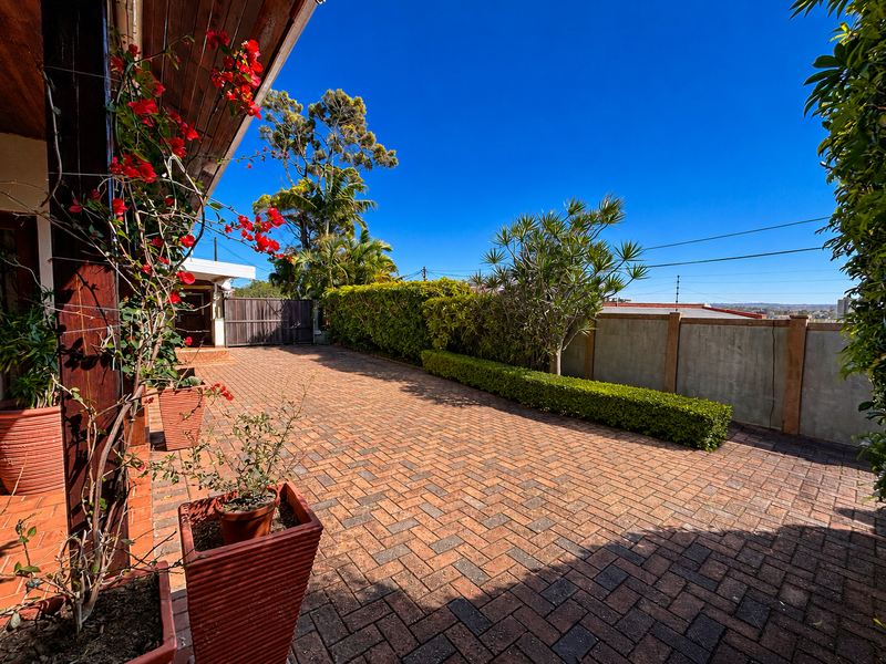 A well-maintained brick-paved front driveway and entrance area featuring a timber roof overhang, established garden hedging, and a secure concrete boundary wall.